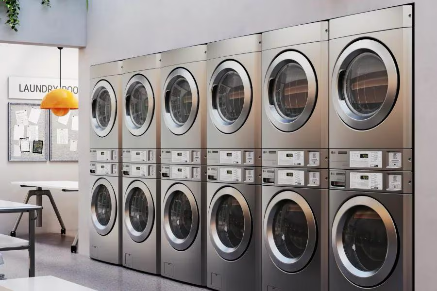 Stacked commercial washer-dryers in a bright communal laundry room with folding tables and a pendant light