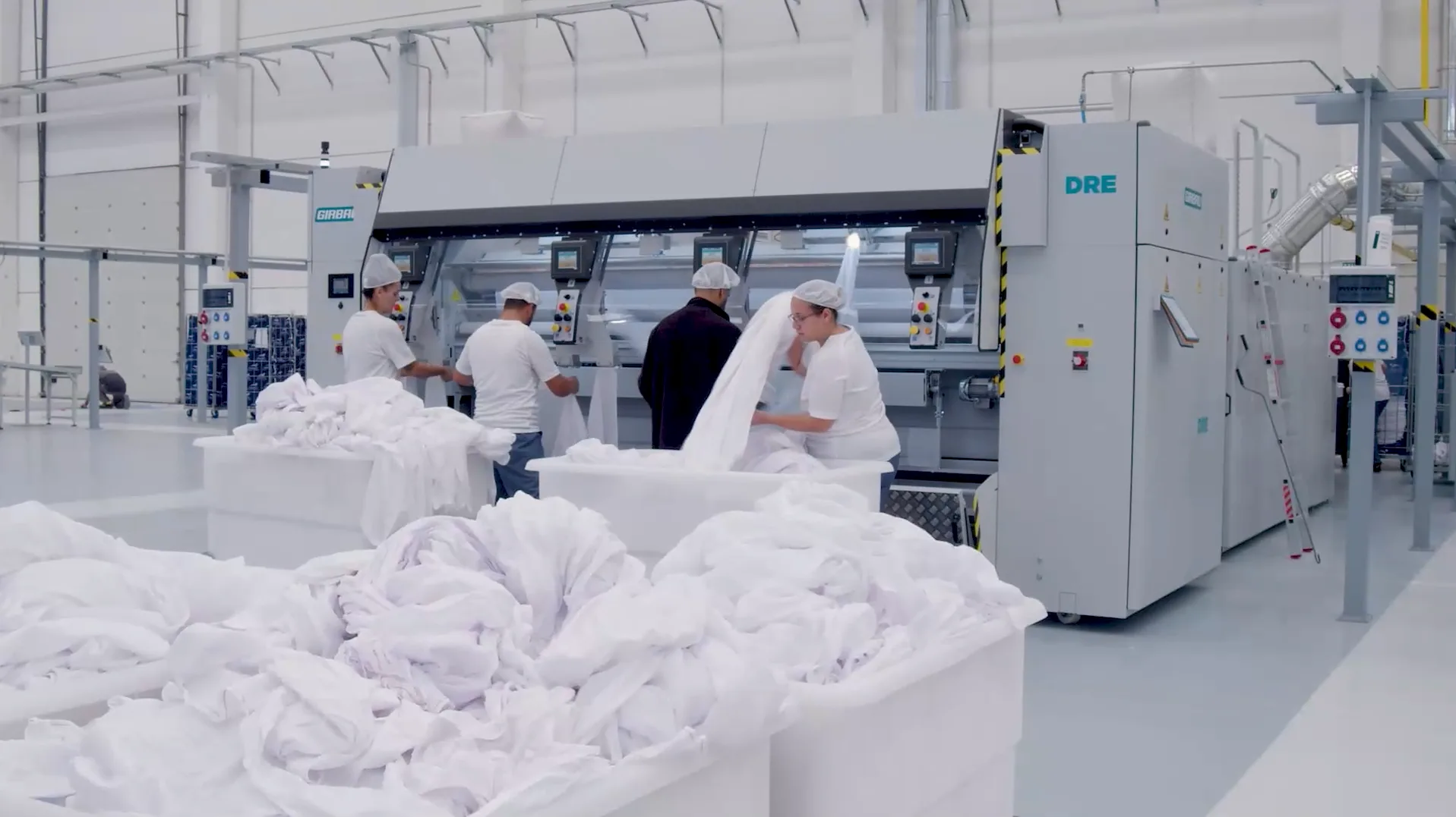Girbau DRE flatwork ironer line in a commercial laundry, with four workers in hairnets feeding white linens and tubs of clean sheets in the foreground