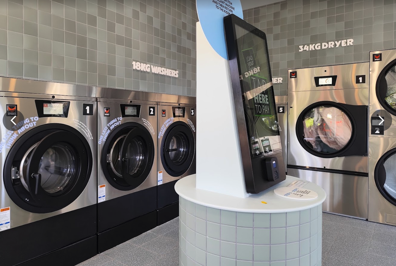 A cashless payment kiosk between rows of washers and dryers in a modern laundromat