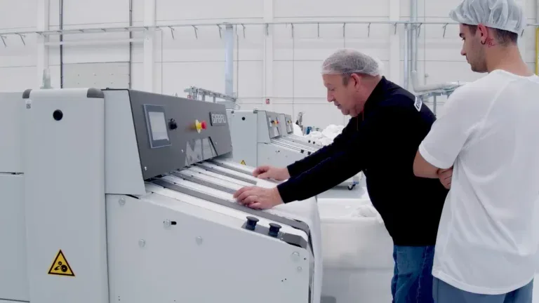 Girbau flatwork ironer line in a commercial laundry, with two workers in hairnets feeding linen